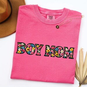 May include: A pink t-shirt with the words "BOY MOM" in a floral design. The shirt has a crew neck and a tag that says "Comfort Colors". A gold necklace with a small pendant is on the shirt.