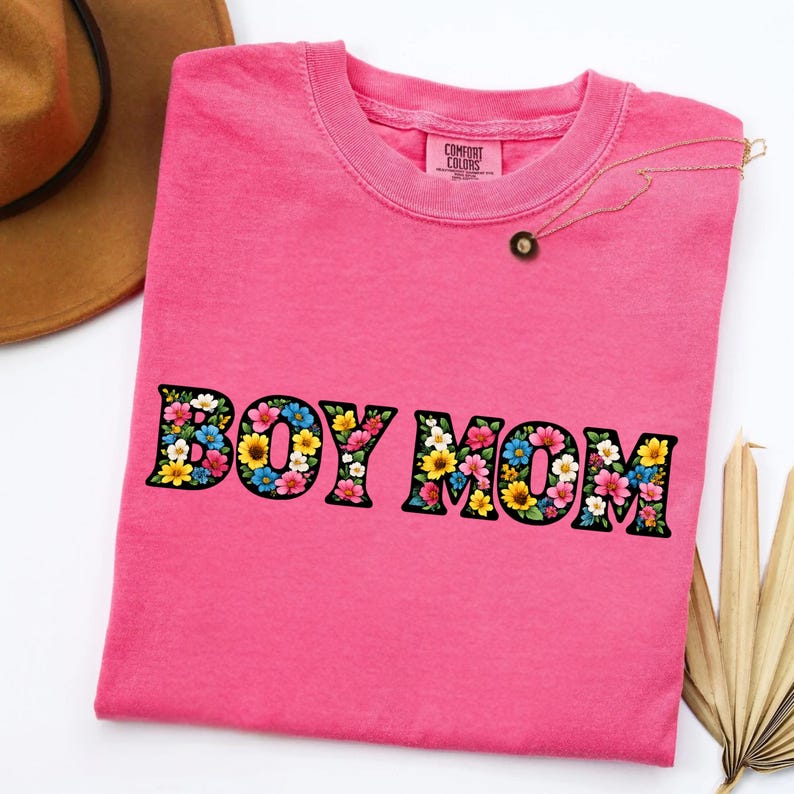 May include: A pink t-shirt with the words "BOY MOM" in a floral design. The shirt has a crew neck and a tag that says "Comfort Colors". A gold necklace with a small pendant is on the shirt.