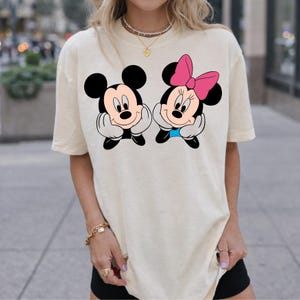 May include: Cream-colored t-shirt featuring a graphic of Mickey Mouse and Minnie Mouse. Mickey is wearing black shorts and has a smiling expression. Minnie has a pink bow and a blue bow tie. Both characters are resting their chins on their hands.