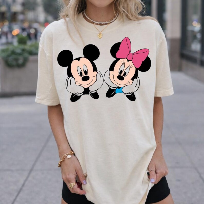 May include: Cream-colored t-shirt featuring a graphic of Mickey Mouse and Minnie Mouse. Mickey is wearing black shorts and has a smiling expression. Minnie has a pink bow and a blue bow tie. Both characters are resting their chins on their hands.