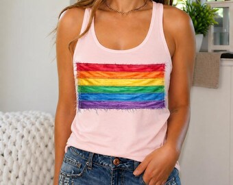May include: A light pink tank top featuring a rectangular patch with horizontal stripes in red, orange, yellow, green, blue, and purple. The patch has a frayed, raw-edge texture. The tank top has a scoop neckline and wide shoulder straps.