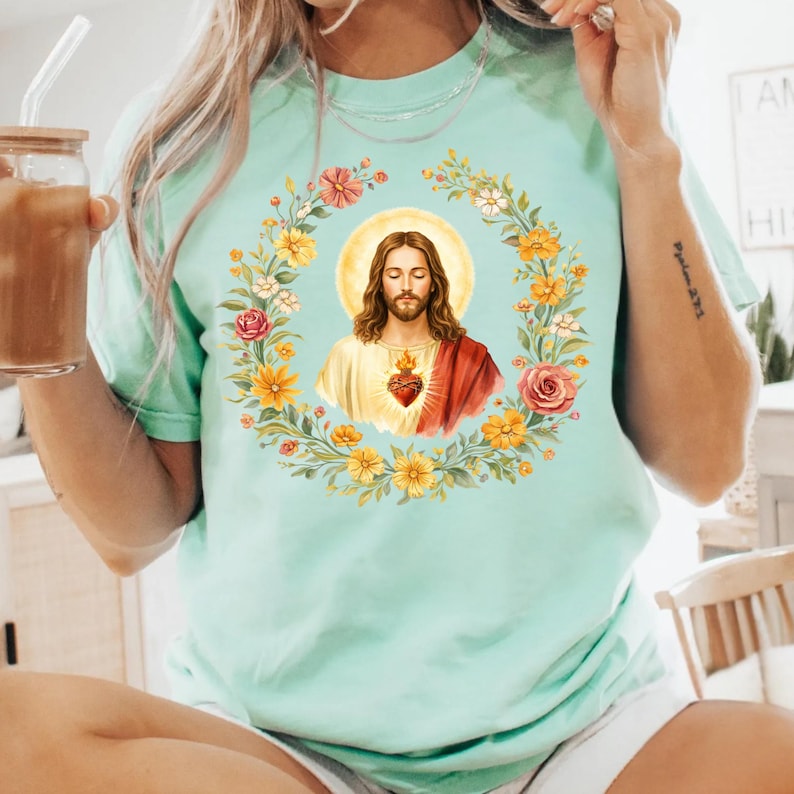 K&ouml;nnte beinhalten: Ein hellgr&uuml;nes T-Shirt mit einem floralen Kranzdesign und einem Portr&auml;t von Jesus Christus. Das Portr&auml;t zeigt Jesus mit langen braunen Haaren, einem roten Gewand und einem Herzsymbol. Der Kranz enth&auml;lt gelbe, rosa und orangefarbene Blumen.