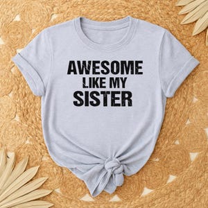 May include: A light gray t-shirt with the text "AWESOME LIKE MY SISTER" in bold black letters. The shirt has short sleeves and is knotted at the bottom. The shirt is displayed on a woven, tan surface.