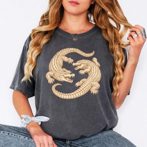 May include: Dark grey t-shirt featuring a detailed illustration of two facing alligators in a circular design. The alligators are in a light tan color, with visible scales and open mouths. The shirt is a casual style.