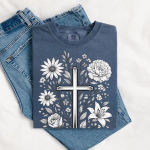 Op de afbeelding: Een marineblauw T-shirt met een wit kruisontwerp omringd door witte bloemen en bladeren. Het T-shirt ligt op een blauwe spijkerbroek.