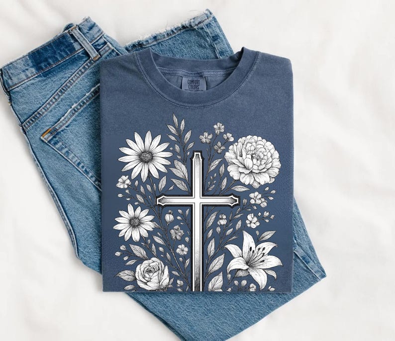 Op de afbeelding: Een marineblauw T-shirt met een wit kruisontwerp omringd door witte bloemen en bladeren. Het T-shirt ligt op een blauwe spijkerbroek.