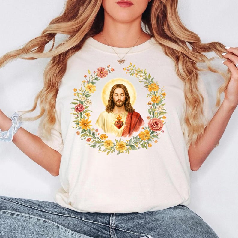 K&ouml;nnte beinhalten: Cremefarbenes T-Shirt mit einer religi&ouml;sen Illustration. Das Design zeigt ein Portr&auml;t von Jesus in einem Blumenkranz, mit goldenem Heiligenschein und einem Herzsymbol. Das Shirt besteht aus einem weichen, bequemen Material.