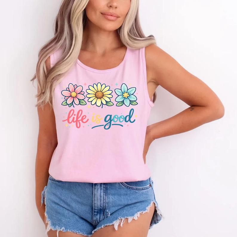 May include: Pink tank top with a floral design and the words "life is good" in colorful script. The top features three flowers in pink, yellow, and blue. The model is wearing denim shorts.