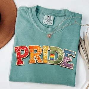 May include: A teal short-sleeve t-shirt with a crew neckline and a Comfort Colors label. The front features the word PRIDE in large, block-style letters, each filled with a different colorful fabric pattern including floral, plaid, and rainbow prints.