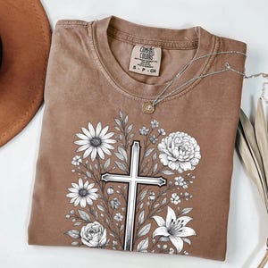 Op de afbeelding: Een bruin Comfort Colors T-shirt met een bloemenkruisontwerp. De afbeelding bevat een zilveren kruis omringd door witte bloemen en bladeren. Een zilveren ketting met een hanger ligt op het shirt.
