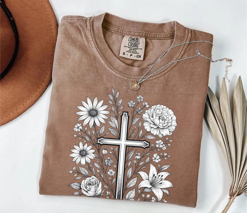 Op de afbeelding: Een bruin Comfort Colors T-shirt met een bloemenkruisontwerp. De afbeelding bevat een zilveren kruis omringd door witte bloemen en bladeren. Een zilveren ketting met een hanger ligt op het shirt.