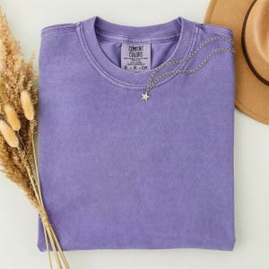 May include: A solid purple short-sleeve t-shirt with a crew neckline and a Comfort Colors brand label printed on the inside collar. A thin silver-toned chain necklace with a small star-shaped pendant rests on the fabric of the shirt.