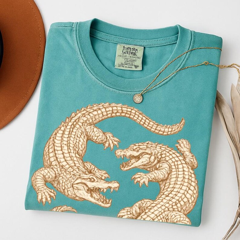 May include: Teal-colored t-shirt featuring a detailed illustration of two alligators facing each other. The shirt has a crew neck and a small tag. A gold necklace with a pendant is draped across the shirt.
