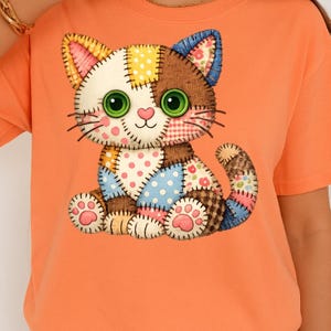 May include: Orange t-shirt featuring a patchwork cat design. The cat has green eyes, pink cheeks, and a variety of fabric patterns in its design, including polka dots, floral prints, and plaid. The cat is sitting with its paws visible.