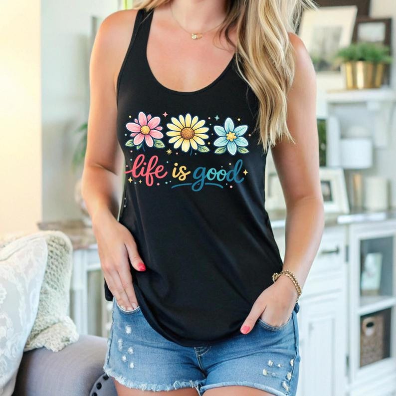 May include: Black tank top with a floral design and the phrase "life is good" in colorful script. The top features three flowers in pink, yellow, and blue. The model is wearing denim shorts.