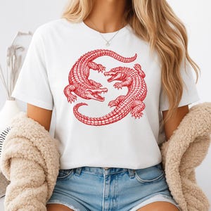 May include: White t-shirt featuring a red illustration of two alligators in a circular design. The alligators are detailed with scales and open mouths. The shirt is made of a soft material and has short sleeves.