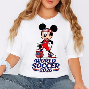 May include: White t-shirt featuring a cartoon Mickey Mouse in a red soccer uniform with a US flag headband. Mickey is standing on a soccer ball with a US flag design. The text "WORLD SOCCER 2026" is below the image, with red and blue stripes.