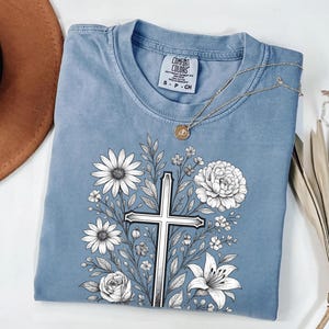 Op de afbeelding: Een blauw Comfort Colors T-shirt met een bloemenkruisontwerp. De afbeelding bevat een zilveren kruis omringd door witte bloemen en groene bladeren. Een gouden ketting met een hanger is over het shirt gedrapeerd.