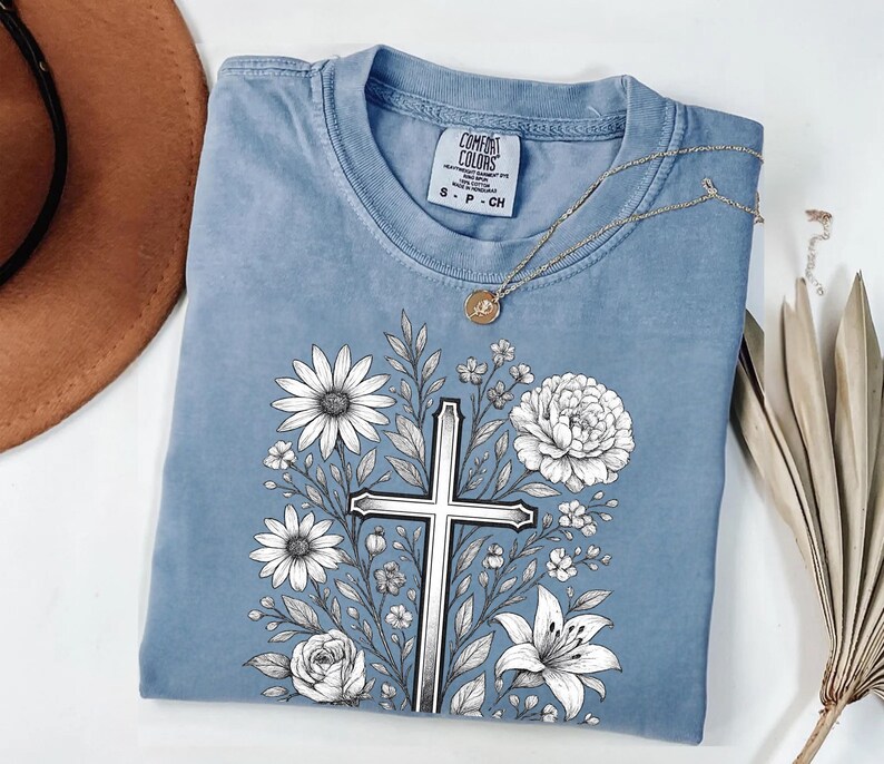 Op de afbeelding: Een blauw Comfort Colors T-shirt met een bloemenkruisontwerp. De afbeelding bevat een zilveren kruis omringd door witte bloemen en groene bladeren. Een gouden ketting met een hanger is over het shirt gedrapeerd.