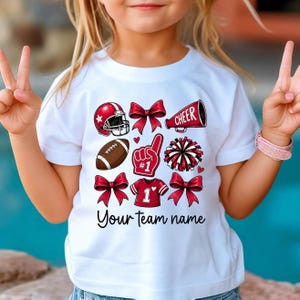 May include: A white short-sleeve t-shirt featuring a graphic print of a red football helmet, a brown football, a red megaphone with the word CHEER, a red foam finger with the number 1, a pom-pom, a jersey with the number 1, and red bows.
