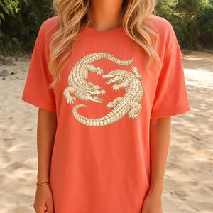 May include: Coral-colored t-shirt featuring a detailed illustration of two alligators in a circular design. The alligators are depicted in a cream color with intricate scale patterns, set against the solid-colored tee. The shirt has short sleeves and a relaxed fit.