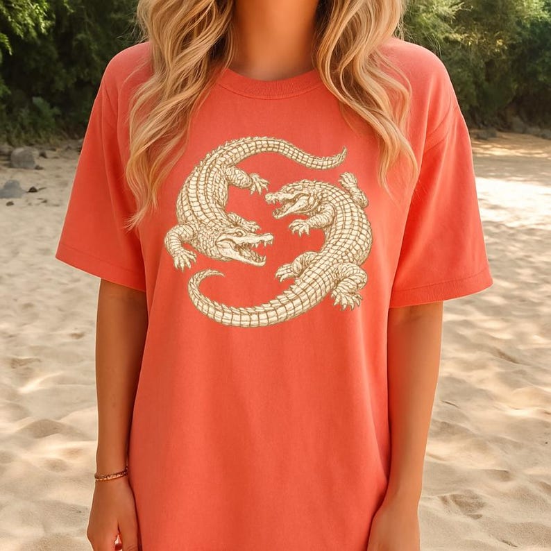 May include: Coral-colored t-shirt featuring a detailed illustration of two alligators in a circular design. The alligators are depicted in a cream color with intricate scale patterns, set against the solid-colored tee. The shirt has short sleeves and a relaxed fit.