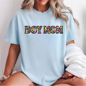May include: A light blue t-shirt with the text "BOY MOM" in a floral design. The letters are filled with colorful flowers, including yellow, pink, and blue blooms. The shirt has short sleeves and a crew neck.