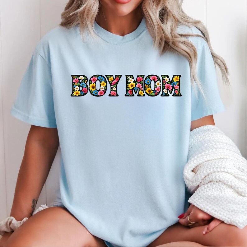 May include: A light blue t-shirt with the text "BOY MOM" in a floral design. The letters are filled with colorful flowers, including yellow, pink, and blue blooms. The shirt has short sleeves and a crew neck.