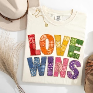 May include: A cream-colored t-shirt featuring the text LOVE WINS in large, multi-colored, patchwork-style letters. Each letter is composed of various fabric patterns in red, orange, yellow, green, blue, purple, and pink, with visible stitching.