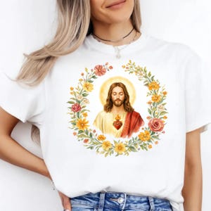 K&ouml;nnte beinhalten: Wei&szlig;es T-Shirt mit einer Aquarellillustration von Jesus, umgeben von einem Blumenkranz. Das Bild enth&auml;lt ein Herz mit Flammen. Das Design ist mittig auf dem T-Shirt platziert.