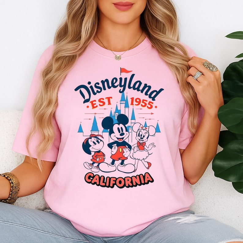 May include: Pale pink t-shirt with a Disneyland graphic. The design features Mickey Mouse, Minnie Mouse, and Donald Duck, with the words "Disneyland EST 1955 CALIFORNIA". The graphic is in a vintage style, with a castle and red flag.