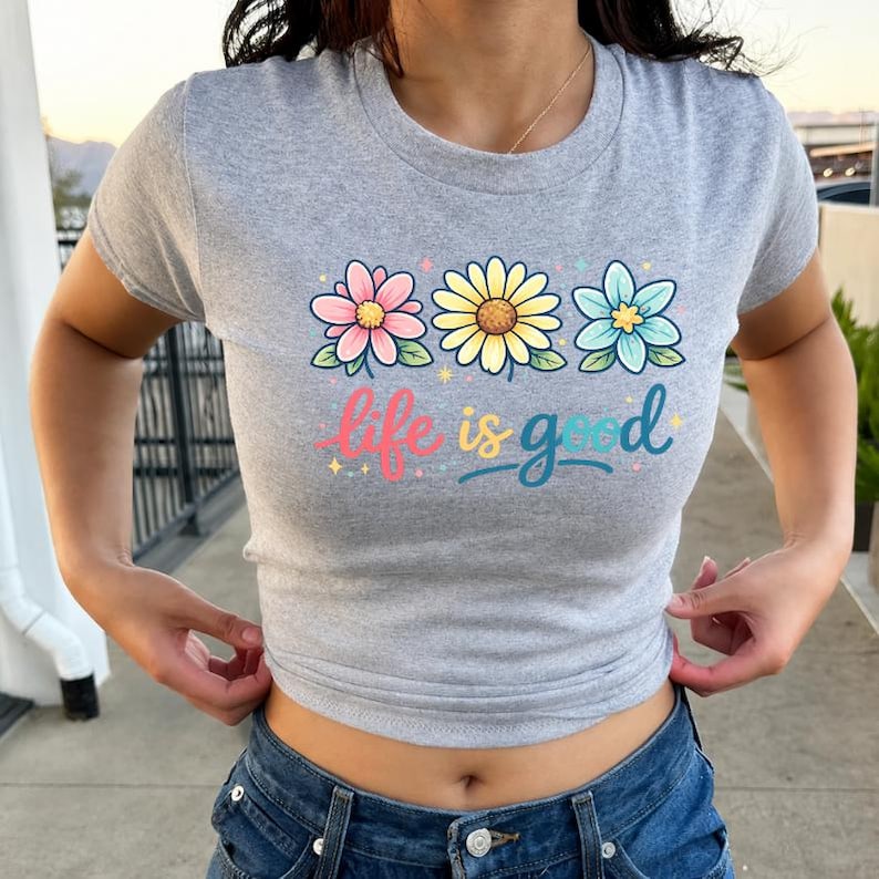 May include: A gray t-shirt with a crew neck featuring a floral design and the phrase "life is good." The graphic includes three colorful flowers in pink, yellow, and blue, with the text in a playful font.