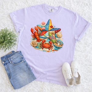 May include: A light purple t-shirt featuring a colorful patchwork starfish, a red lobster, and a red crab. The design includes seashells and a pearl. The shirt is paired with blue jeans and white sneakers, suggesting a casual, beach-themed style.