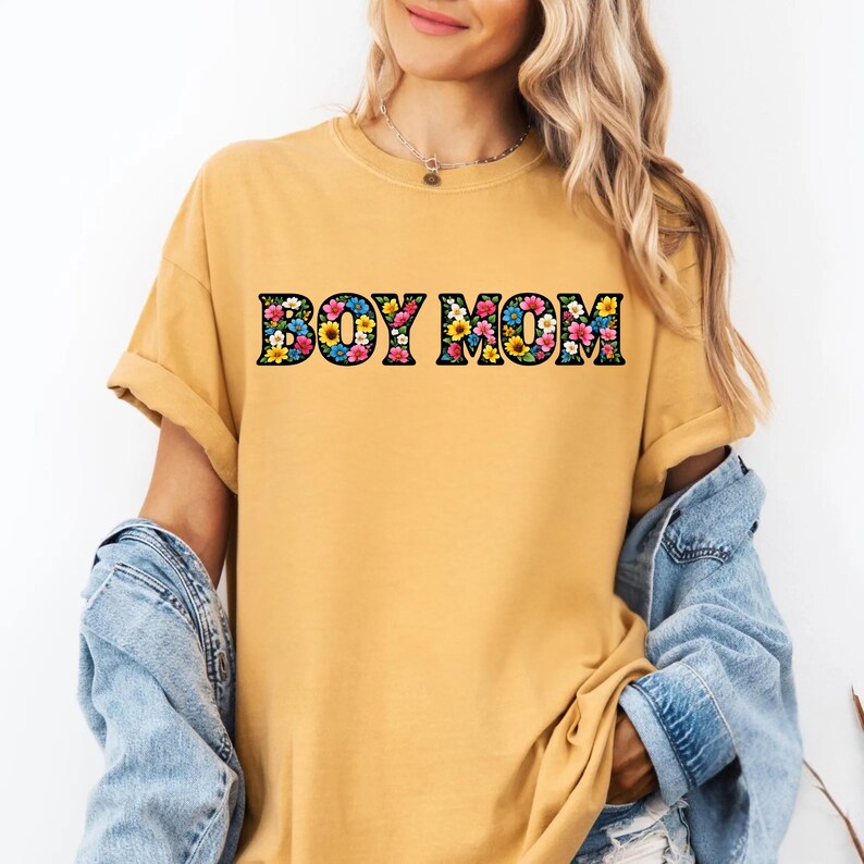 May include: Mustard yellow t-shirt with the text "BOY MOM" in a floral design. The shirt has short sleeves and a relaxed fit. A denim jacket is worn over the shirt.