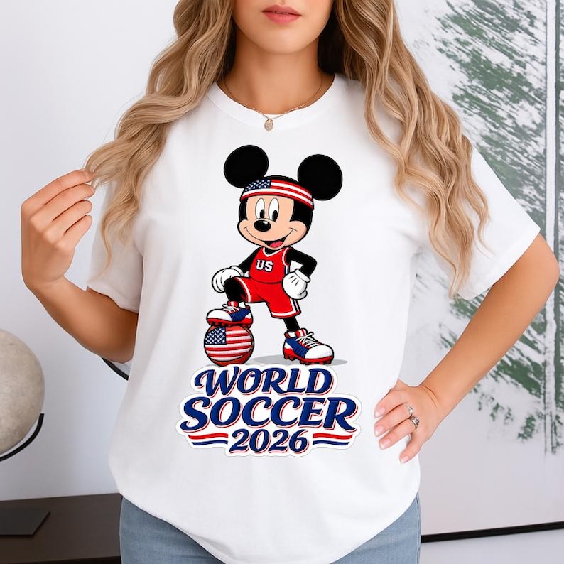 May include: White t-shirt featuring a cartoon Mickey Mouse in a soccer uniform with a US flag headband. The design includes the text "WORLD SOCCER 2026" in blue and red. The character is standing on a soccer ball.