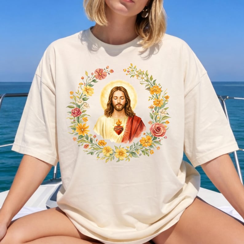 K&ouml;nnte beinhalten: Cremefarbenes T-Shirt mit einer Aquarellillustration von Jesus mit Heiligenschein, umgeben von einem floralen Kranz aus gelben und rosa Blumen. Das Bild hat eine Vintage-&Auml;sthetik.