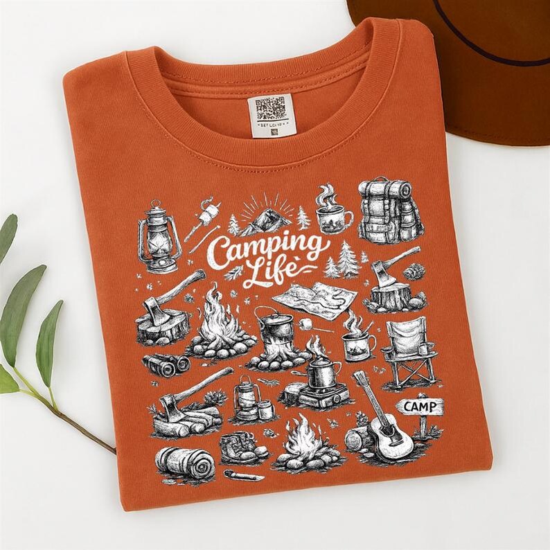 May include: Burnt orange t-shirt with a graphic design featuring camping-related illustrations. The design includes a lantern, campfire, backpack, axe, and the words "Camping Life". A brown hat is visible in the background.