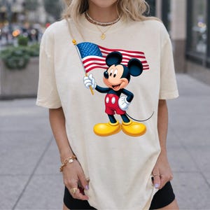 Puede incluir: Camiseta color crema con un gr&aacute;fico de Mickey Mouse sosteniendo una bandera estadounidense. Mickey lleva pantalones cortos rojos, zapatos amarillos y guantes blancos. La bandera tiene rayas rojas y blancas y un campo azul con estrellas blancas.