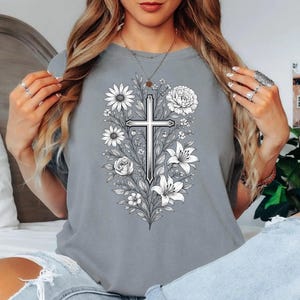 Op de afbeelding: Grijze t-shirt met een gedetailleerd zwart-wit bloemen kruisontwerp. Het ontwerp bevat madeliefjes, rozen en lelies, waardoor een botanisch en religieus thema ontstaat. Het shirt is een comfortabele, casual stijl.