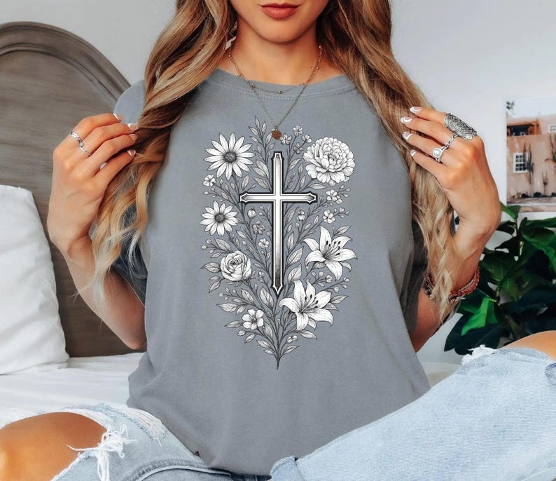 Op de afbeelding: Grijze t-shirt met een gedetailleerd zwart-wit bloemen kruisontwerp. Het ontwerp bevat madeliefjes, rozen en lelies, waardoor een botanisch en religieus thema ontstaat. Het shirt is een comfortabele, casual stijl.