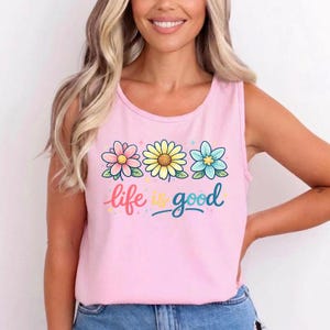 May include: A light pink tank top with a floral design and the words "life is good". The graphic features three flowers in pink, yellow, and blue. The text is in a script font.