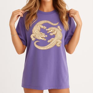 May include: A purple t-shirt featuring a gold-colored illustration of two alligators facing each other. The alligators are detailed with scales and open mouths. The shirt has short sleeves and a relaxed fit.
