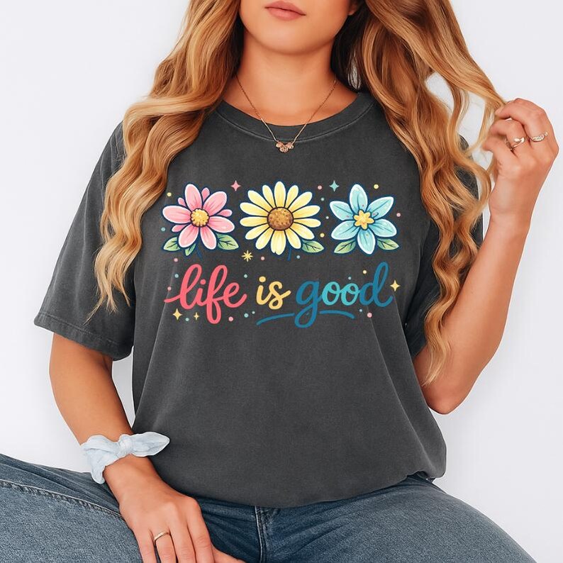May include: Dark gray t-shirt with a floral design and the phrase "life is good." The design features three flowers in pink, yellow, and blue. The text is in a colorful, script font.