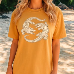 May include: Mustard yellow t-shirt featuring a detailed illustration of two alligators facing each other. The alligators are in a light beige color, with intricate scale and claw details. The shirt has a classic crew neck and short sleeves.