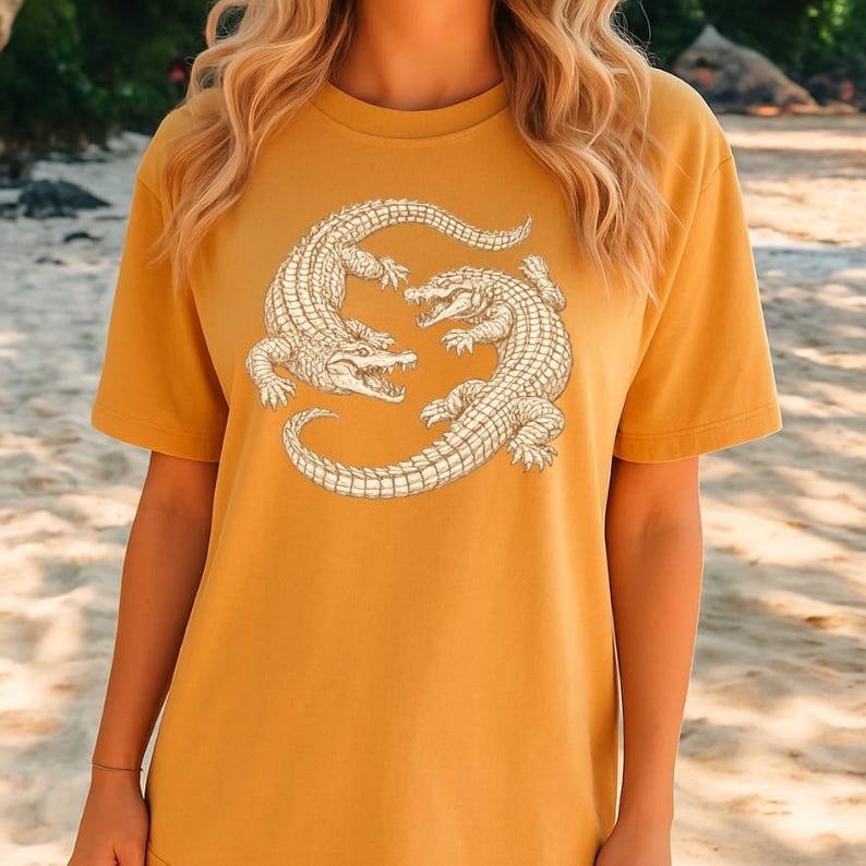 May include: Mustard yellow t-shirt featuring a detailed illustration of two alligators facing each other. The alligators are in a light beige color, with intricate scale and claw details. The shirt has a classic crew neck and short sleeves.