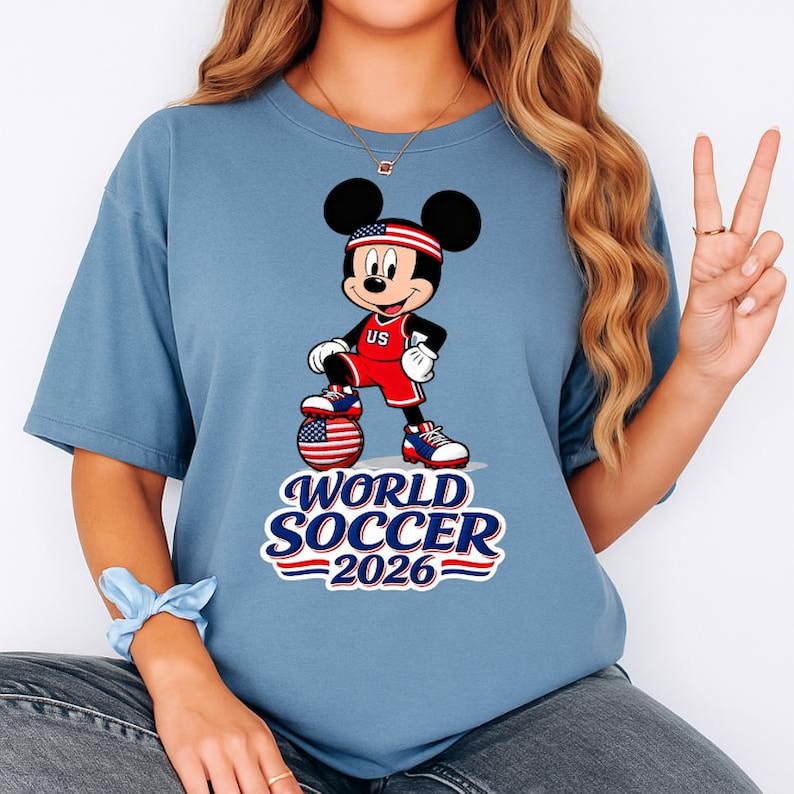 May include: A light blue t-shirt featuring a cartoon Mickey Mouse in a soccer uniform with a soccer ball. The design includes the text "WORLD SOCCER 2026" in red, white, and blue. Mickey is wearing a headband with the US flag.