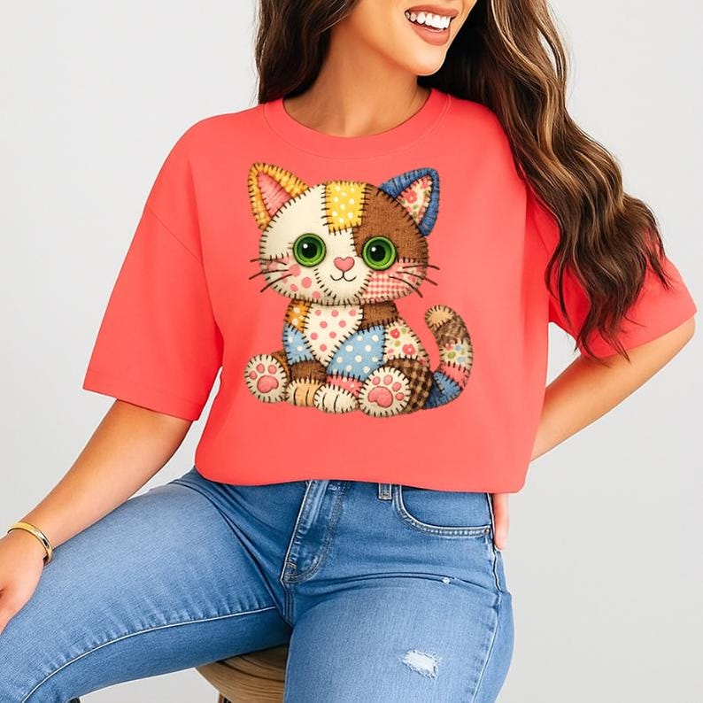 May include: Coral-colored t-shirt featuring a patchwork cat design. The cat has green eyes, a pink nose, and a variety of patterned patches in shades of blue, brown, yellow, and white. The shirt has short sleeves and a relaxed fit.