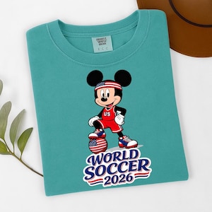 May include: Teal crew neck t-shirt featuring a graphic of Mickey Mouse in a soccer uniform with the text "WORLD SOCCER 2026". Mickey is wearing a red and white uniform with "US" on the chest and a headband with the American flag.