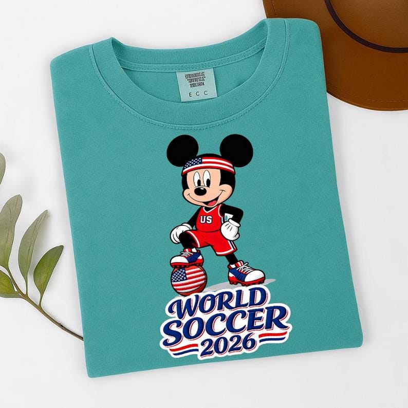 May include: Teal crew neck t-shirt featuring a graphic of Mickey Mouse in a soccer uniform with the text "WORLD SOCCER 2026". Mickey is wearing a red and white uniform with "US" on the chest and a headband with the American flag.