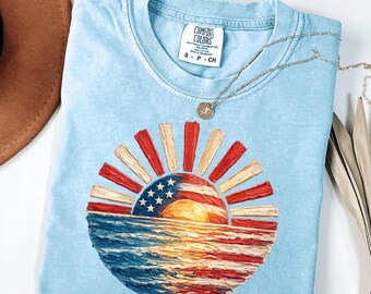May include: A light blue short-sleeve t-shirt with a Comfort Colors label in the neck. The graphic features a sun rising over water, rendered in a textured paint style with red, white, and blue colors resembling the American flag pattern.
